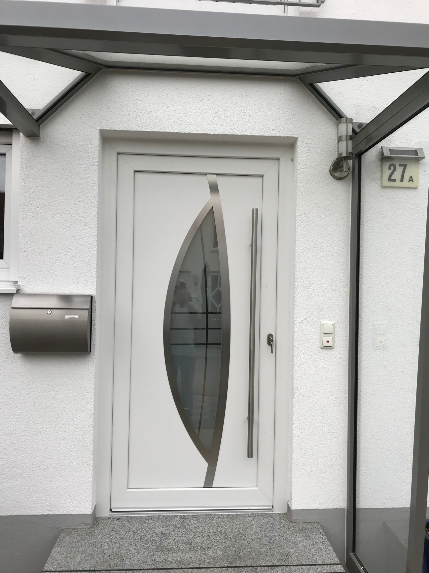 Nowoczesne, białe drzwi wejściowe z ozdobnym przeszkleniem w kształcie łzy, zamontowane w białym budynku z szarą skrzynką na listy i zadaszeniem z metalową konstrukcją.