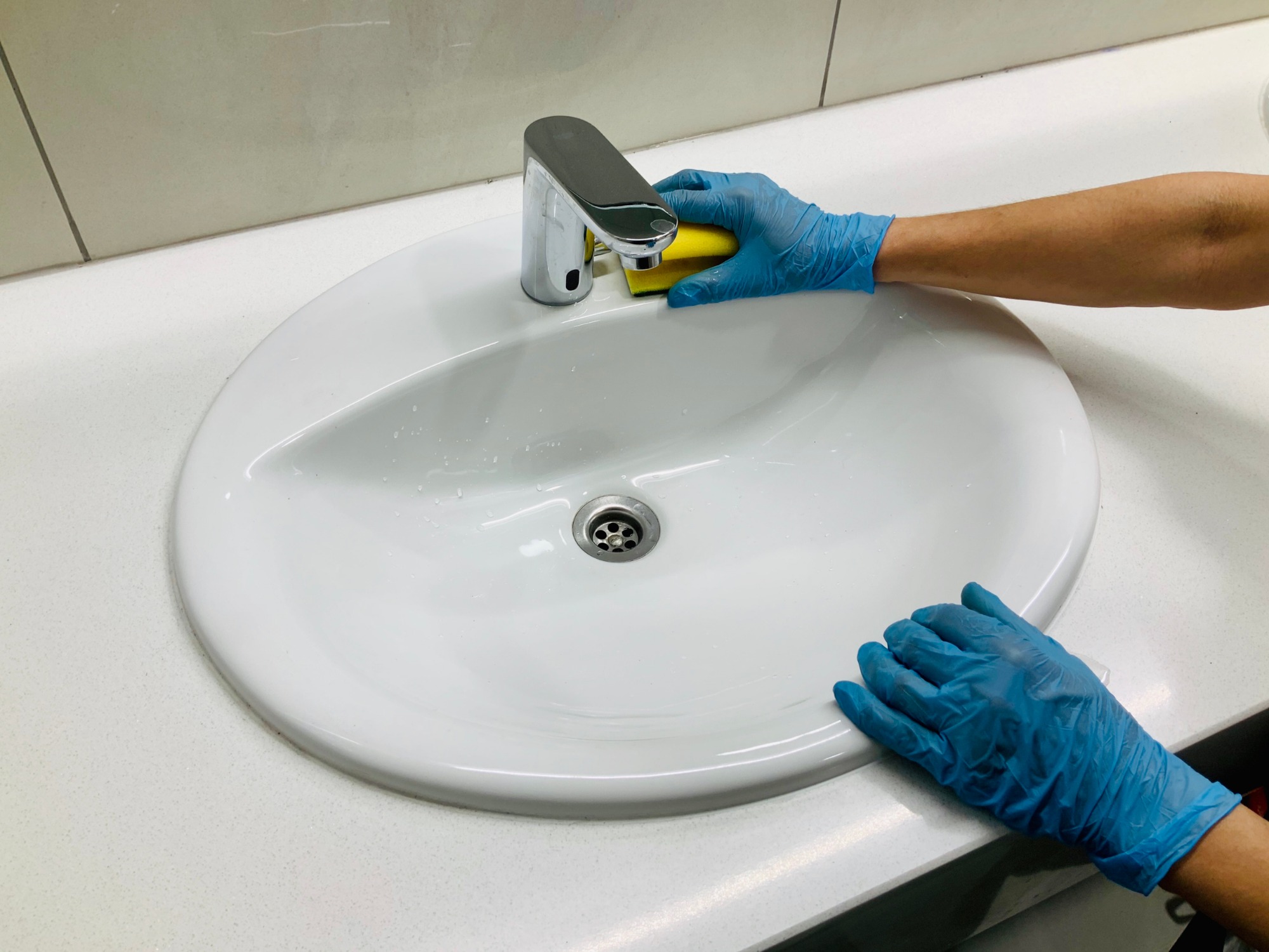Osoba w niebieskich rękawiczkach czyści białą umywalkę z chromowaną baterią za pomocą żółtej gąbki. Widoczne krople wody w umywalce i na blacie.
