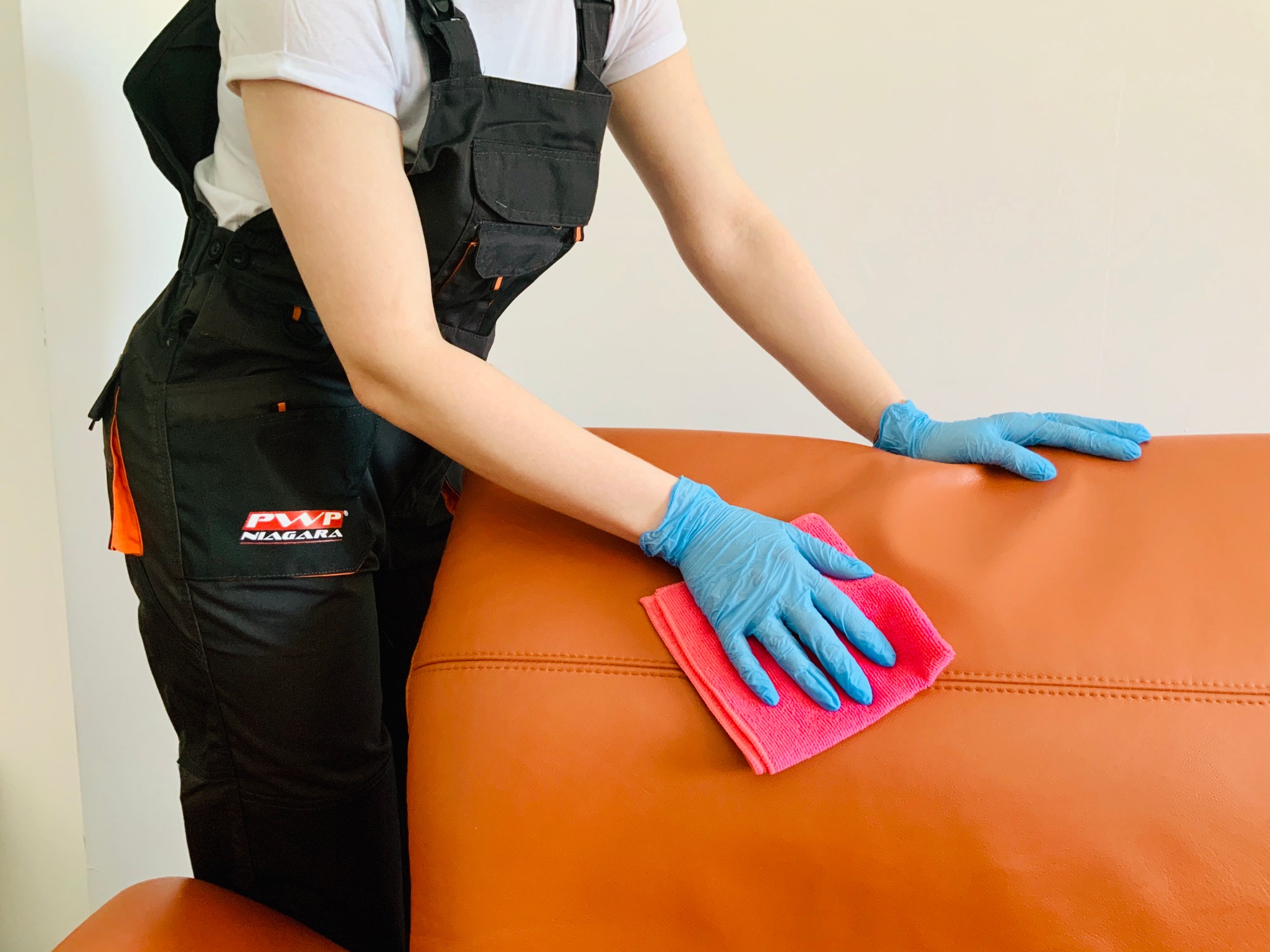 Osoba w kombinezonie PWP Niagara i niebieskich rękawiczkach czyści pomarańczową skórzaną sofę różową ściereczką z mikrofibry.