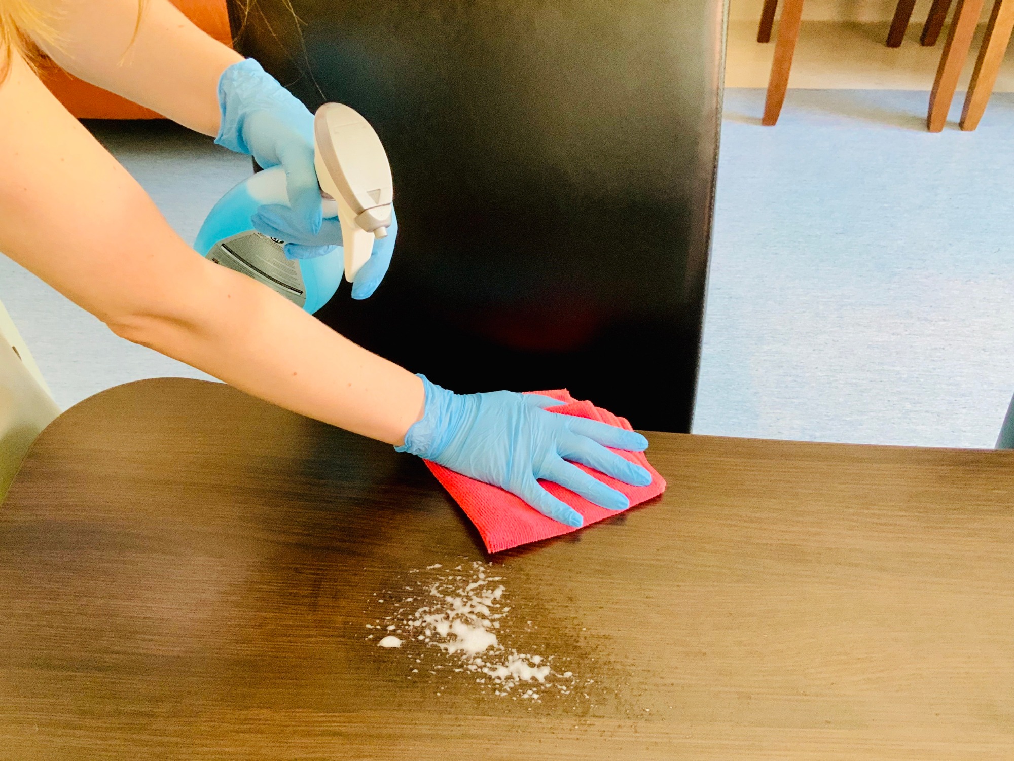 Osoba w niebieskich rękawiczkach spryskuje i czyści brązowy stół czerwoną ściereczką z mikrofibry, widoczne pozostałości po czyszczeniu.