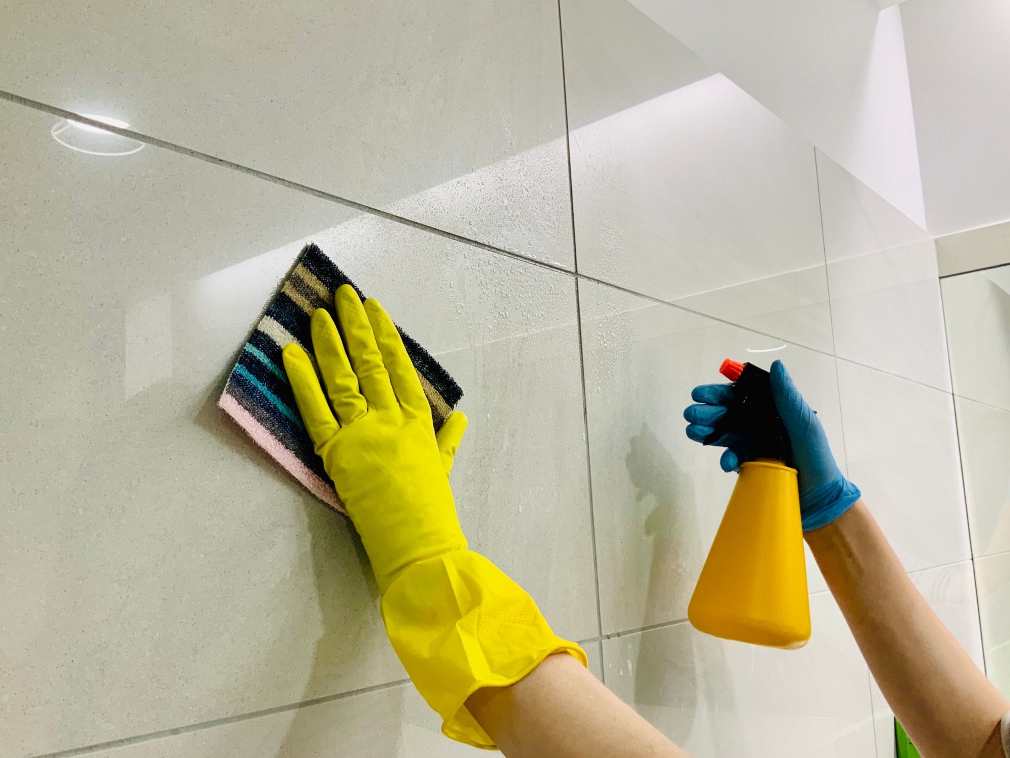 Ręce w żółtych i niebieskich rękawicach czyszczą jasne, kwadratowe płytki ścienne za pomocą kolorowej ściereczki i żółtego spryskiwacza.