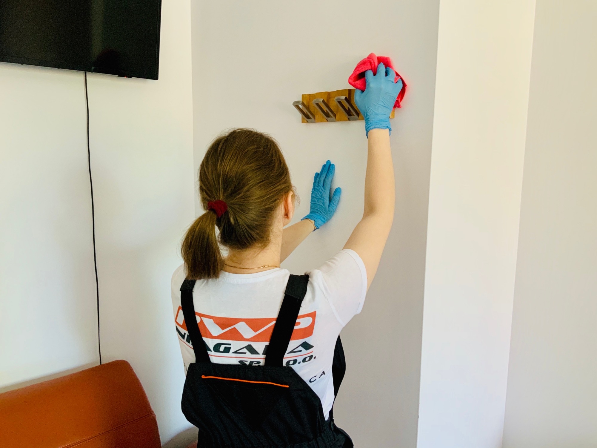 Pracownica sprzątająca ścianę w mieszkaniu, w niebieskich rękawiczkach, używa różowej ściereczki do czyszczenia drewnianego wieszaka na ubrania.
