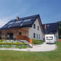 Dom jednorodzinny z zainstalowanymi panelami słonecznymi na dachu, obok zaparkowana biała przyczepa kempingowa, widok z przodu w słoneczny dzień.