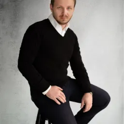 Portret mężczyzny w czarnym swetrze i białej koszuli, siedzącego na stołku na tle szarej ściany, z pierścieniem na palcu i bransoletką na nadgarstku.