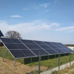 Panel fotowoltaiczny ustawiony na metalowych wspornikach na trawiastym terenie, z widocznym budynkiem w tle i błękitnym niebem z lekkimi chmurami.