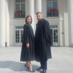 Dwoje prawników w togach, kobieta w szpilkach, mężczyzna z brodą, stoją przed budynkiem z kolumnami i kwiatami w oknach.