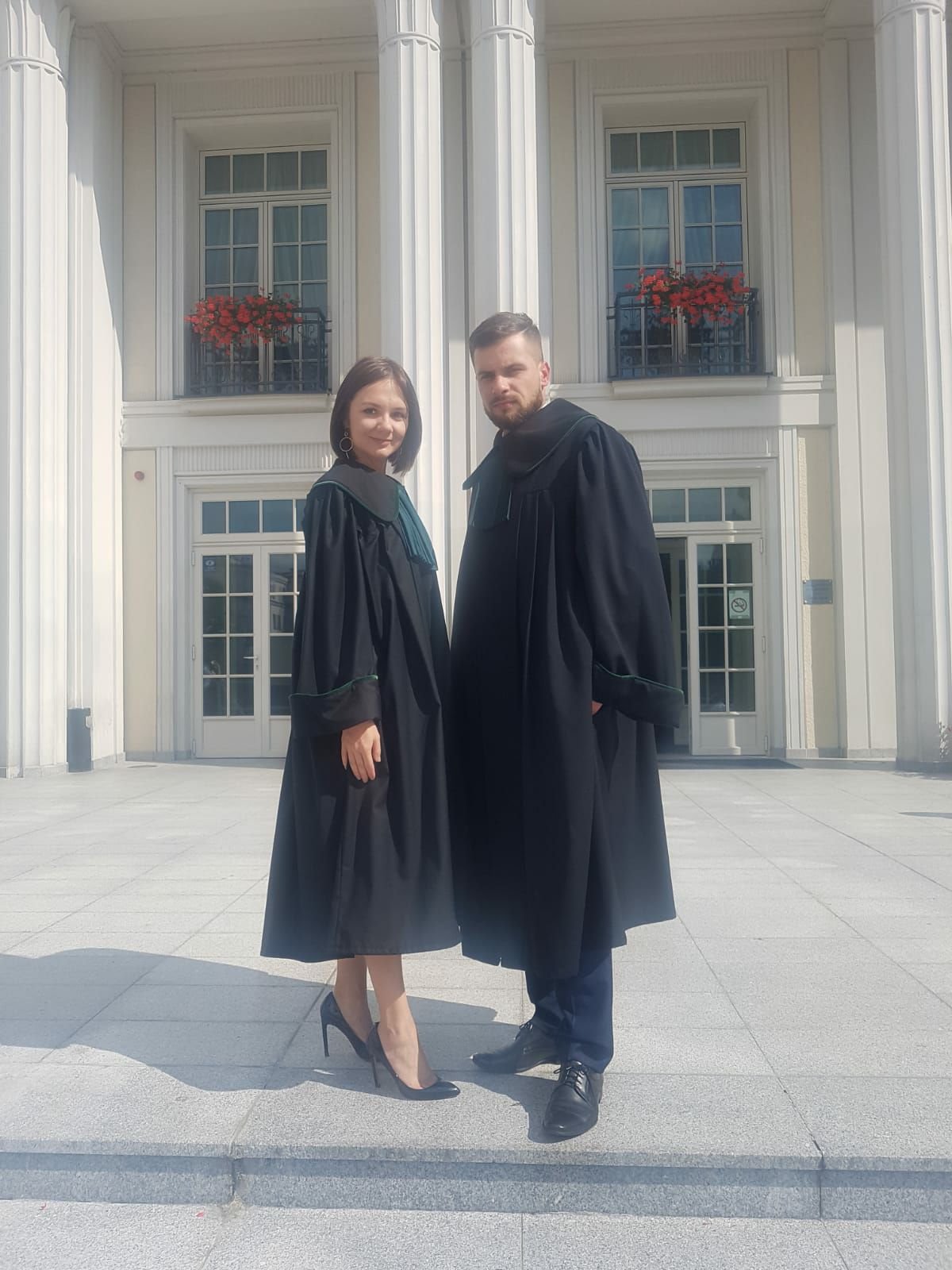 Dwoje prawników w togach, kobieta w szpilkach, mężczyzna z brodą, stoją przed budynkiem z kolumnami i kwiatami w oknach.