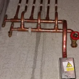 Miedziana instalacja grzewcza z rozdzielaczem i zaworem odcinającym, zamontowana na ścianie z widocznymi rurami zasilającymi pod sufitem, z podłączonym sterownikiem Honeywell z ostrzeżeniem...