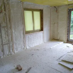 Wnętrze w trakcie prac izolacyjnych: ściany i sufit pokryte warstwą pianki, okno zabezpieczone folią, na podłodze płyty izolacyjne i cegła