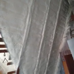 Poddasze z widoczną konstrukcją dachu po natrysku białej piany izolacyjnej, z fragmentami odkrytej cegły i elementami drewnianymi konstrukcji schodów.