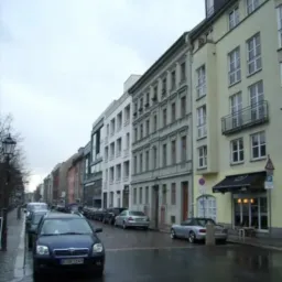 Ulica miejska w pochmurny dzień, z rzędem zaparkowanych samochodów i budynkami o różnej architekturze po obu stronach jezdni. Widoczny szyld nad wejściem do lokalu.