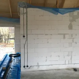 Wewnętrzne okablowanie elektryczne w budynku w trakcie budowy; widoczne puszki instalacyjne, rurki ochronne i wiązka przewodów zwinięta pod sufitem.