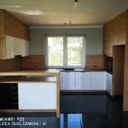 Nowoczesna kuchnia z drewnianymi szafkami górnymi i białymi dolnymi, czarny blat, podłoga z połyskiem, okno z widokiem na zieleń, surowa żarówka na suficie.