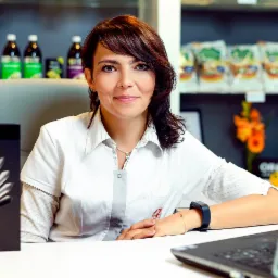 Kobieta w białym fartuchu siedzi za biurkiem z laptopem, za nią półki z produktami zdrowej żywności i napojami.