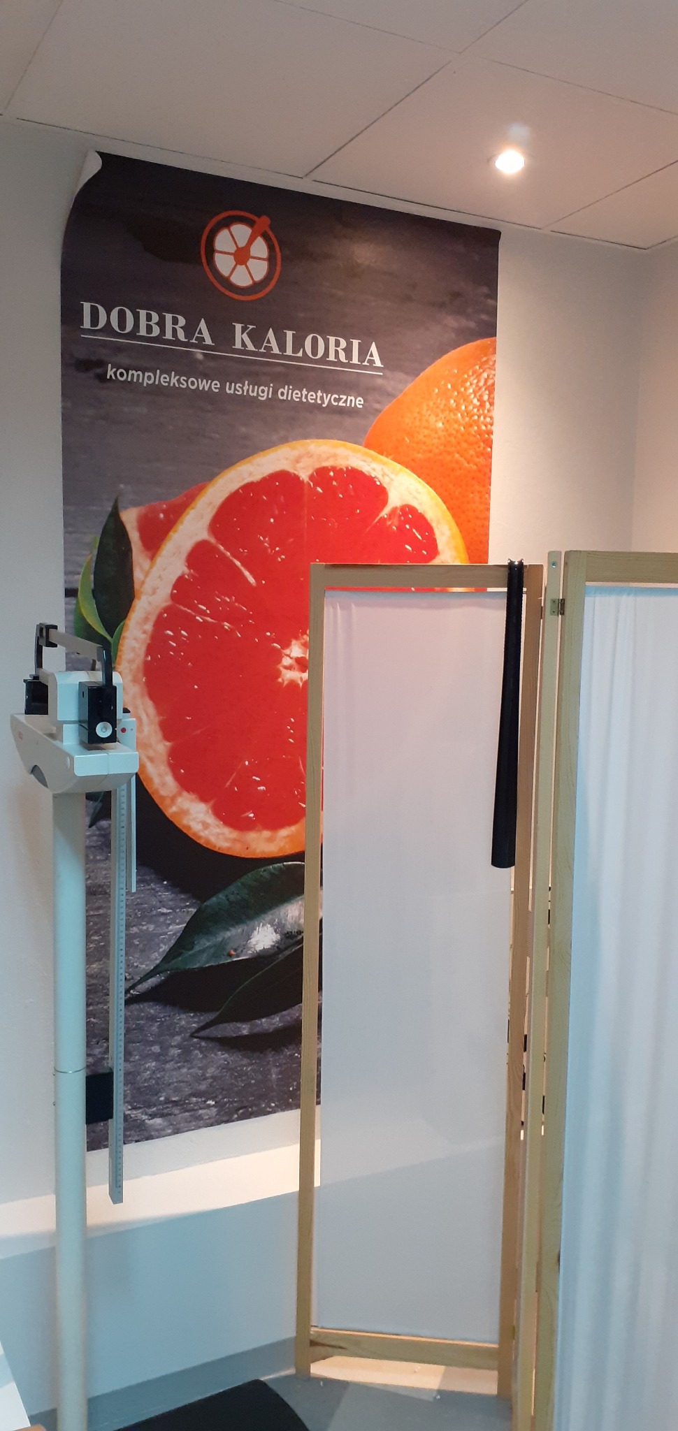 Wnętrze gabinetu z wagą medyczną i parawanem na tle plakatu z przekrojem grejpfruta i napisem 'Dobra Kaloria - kompleksowe usługi ...'