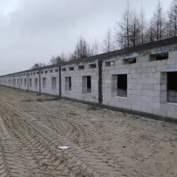 Wznoszenie długiego muru z betonu komórkowego SOLBET z otworami okiennymi, widoczne ślady opon na piaszczystym terenie budowy i drzewa w tle.