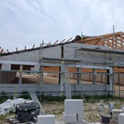 Budowa domu z białych bloczków: widoczne ściany w trakcie wznoszenia, rusztowania, drewniana konstrukcja dachu oraz pracownik na rusztowaniu. Na pierwszym planie agregat prądotwórczy i materiały...