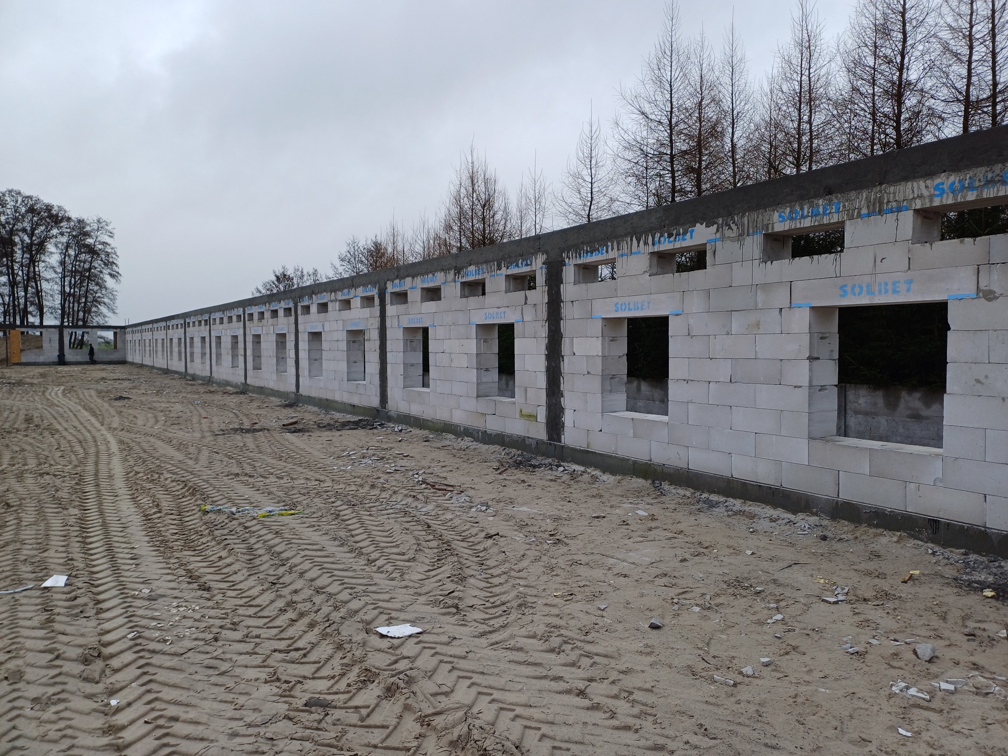 Wznoszenie długiego muru z betonu komórkowego SOLBET z otworami okiennymi, widoczne ślady opon na piaszczystym terenie budowy i drzewa w tle.