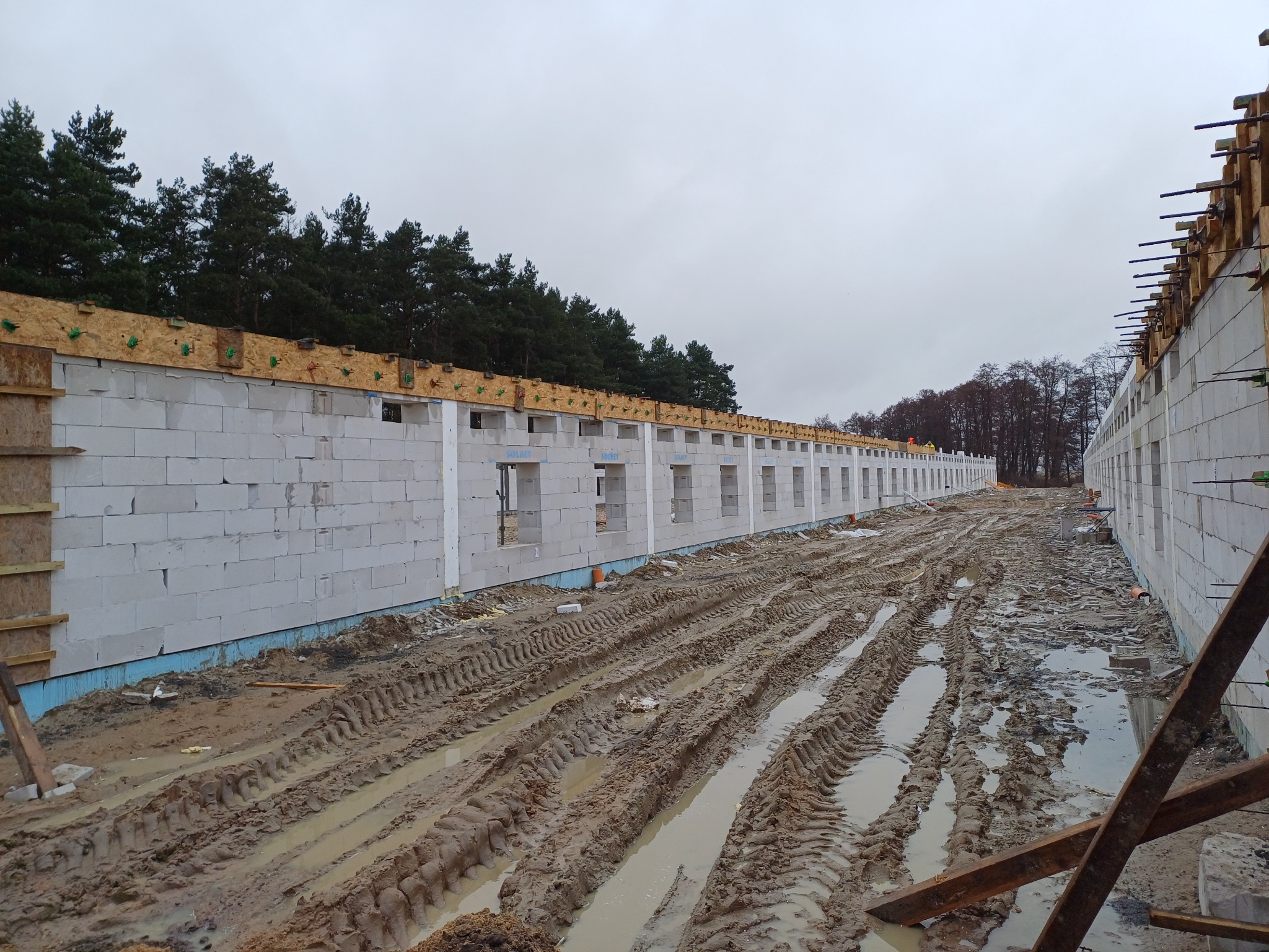 Budowa domu z białych bloczków, widoczne szalunki i zbrojenia, teren budowy z błotnistymi śladami opon, w tle las.