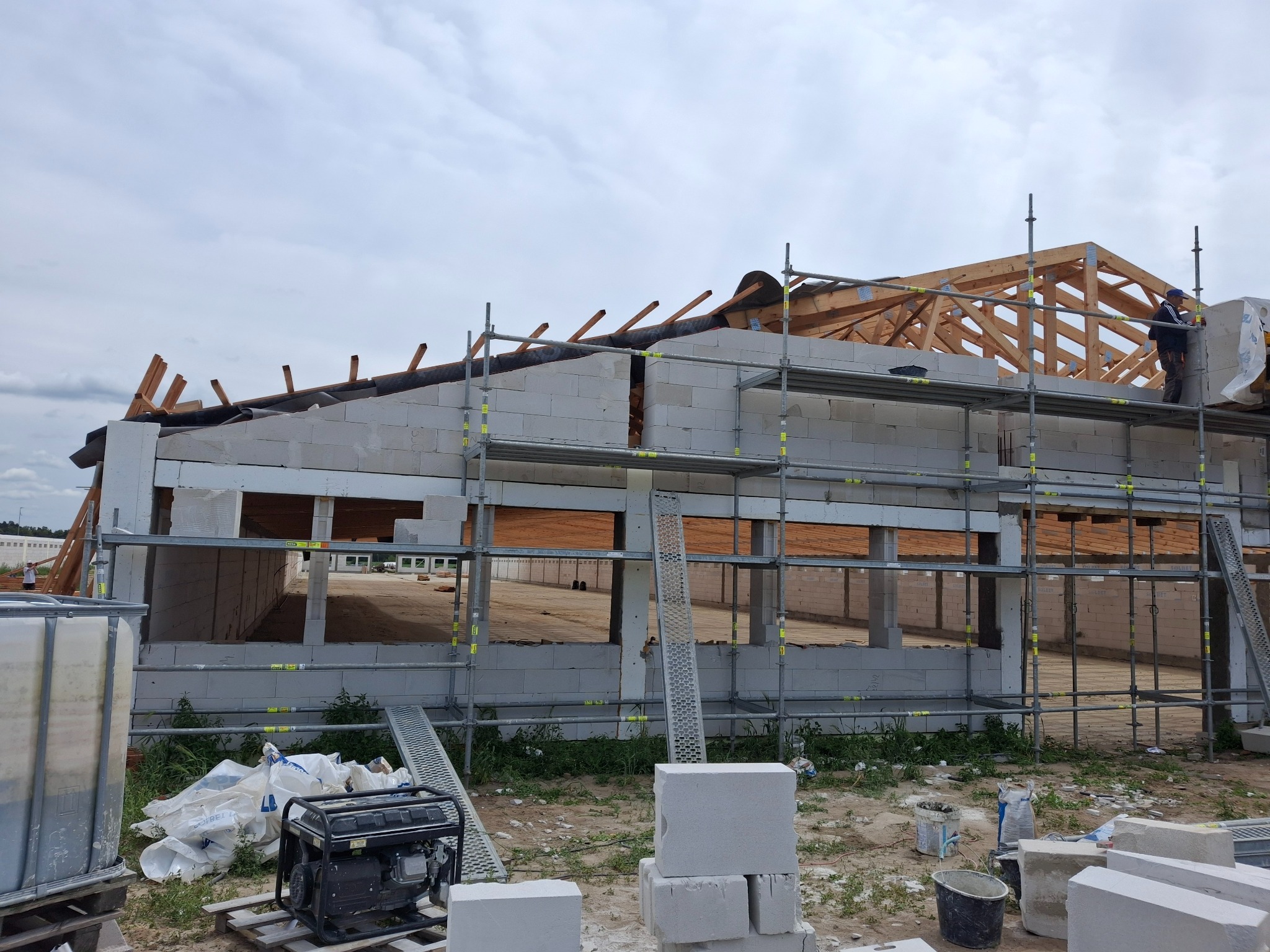 Budowa domu z białych bloczków: widoczne ściany w trakcie wznoszenia, rusztowania, drewniana konstrukcja dachu oraz pracownik na rusztowaniu. Na pierwszym planie agregat prądotwórczy i materiały...