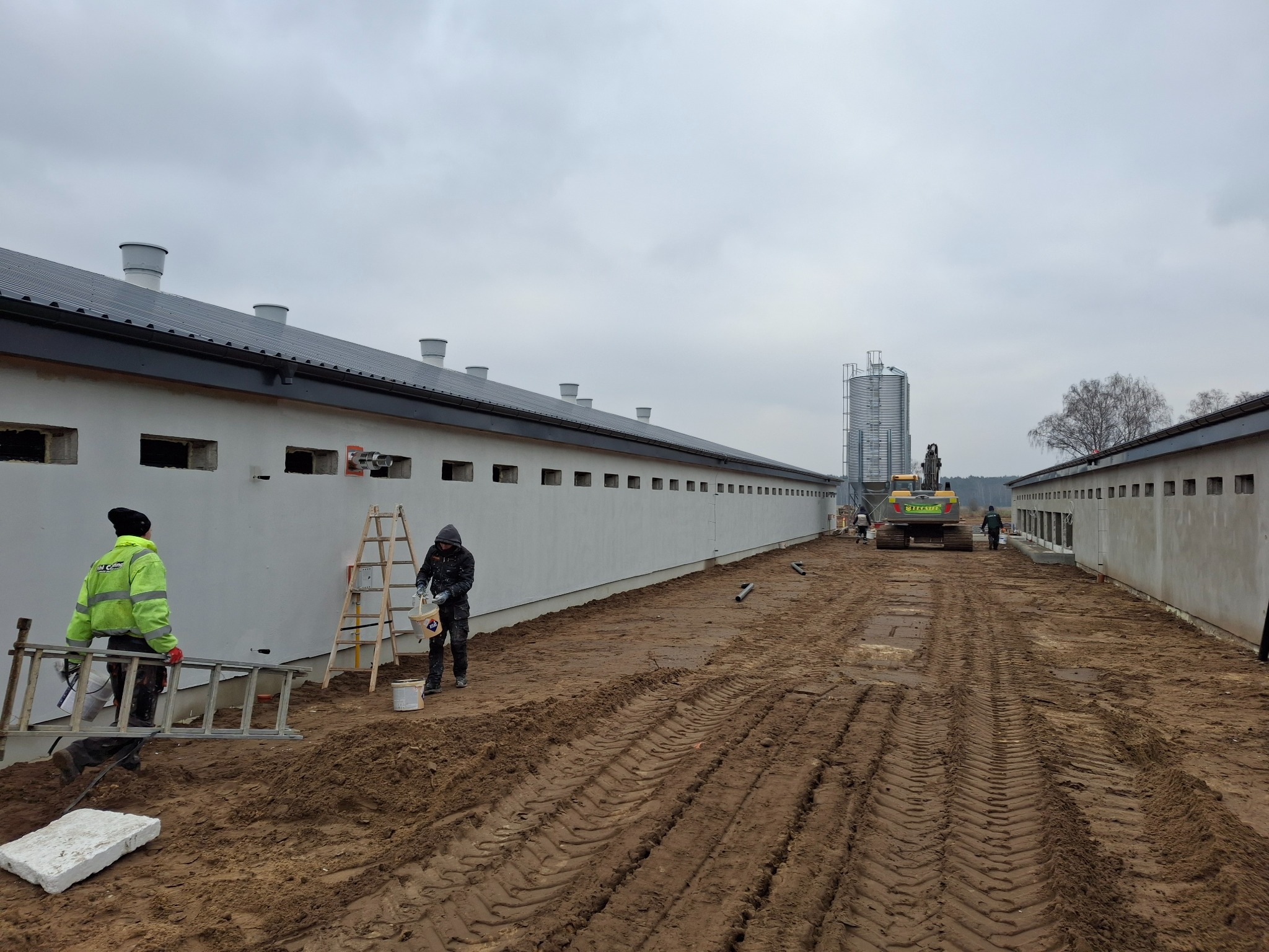 Długi budynek gospodarczy z rzędem okien, świeżo pomalowany na biało, z pracownikami przygotowującymi się do dalszych prac malarskich i sprzętem budowlanym w tle, na placu budowy z błotnistym...