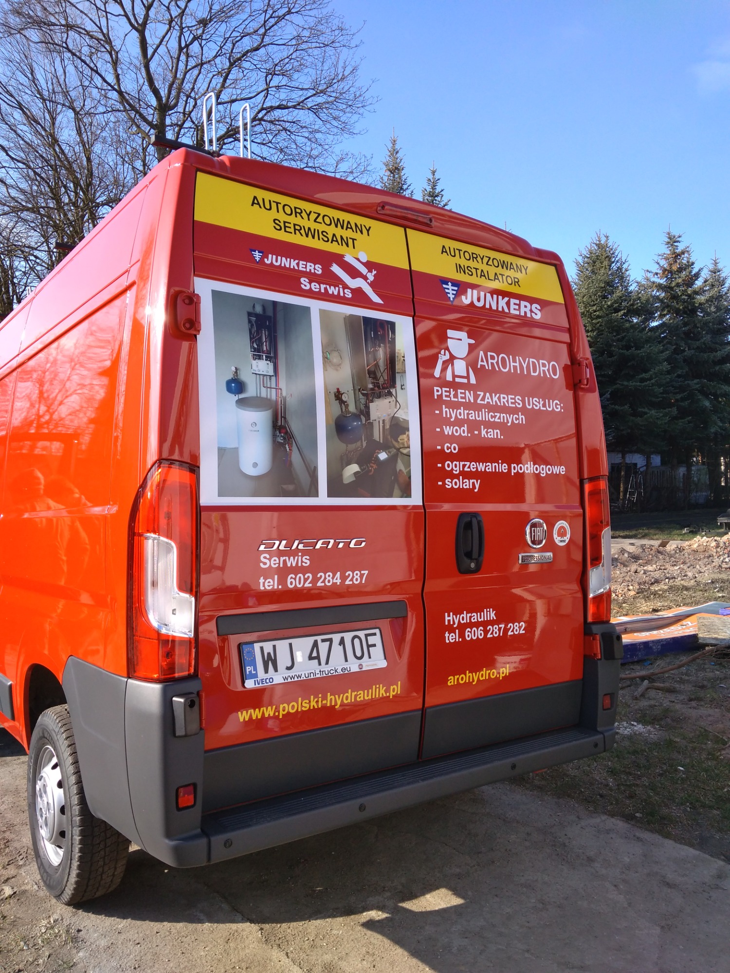 Pomarańczowy samochód dostawczy z oklejeniem reklamowym firmy hydraulicznej Arohydro, prezentujący zdjęcia kotłów i zakres usług: hydraulika, wod-kan, CO, ogrzewanie podłogowe, solary.