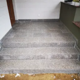 Granitowe schody wejściowe do domu, z widocznymi płytkami i fugami, częściowo ułożone, z ziemią i resztkami materiałów budowlanych u podstawy.