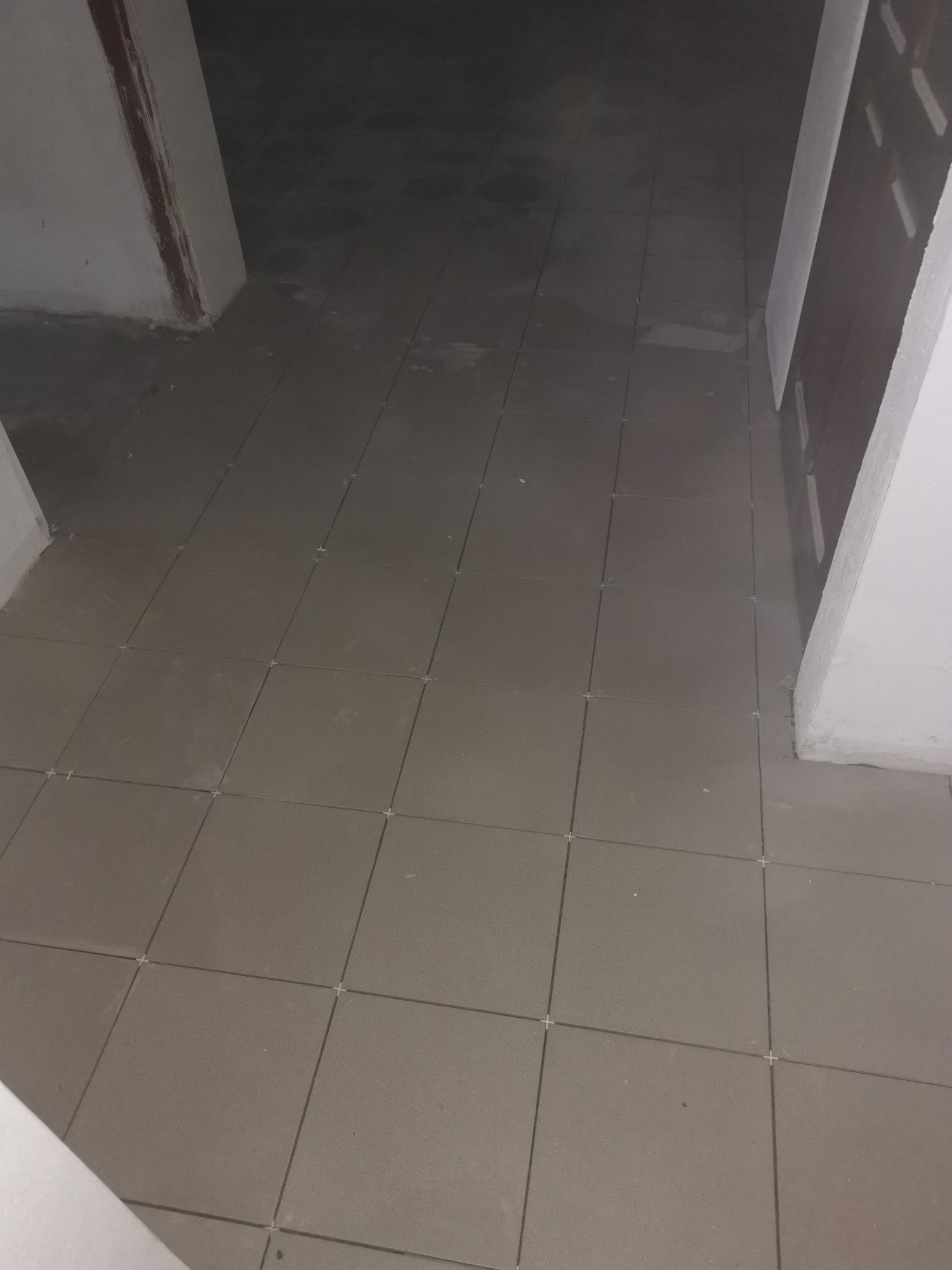 Ułożone szare płytki podłogowe z krzyżykami dystansowymi w trakcie prac wykończeniowych, widoczne fragmenty ścian i ościeżnic w słabym oświetleniu.