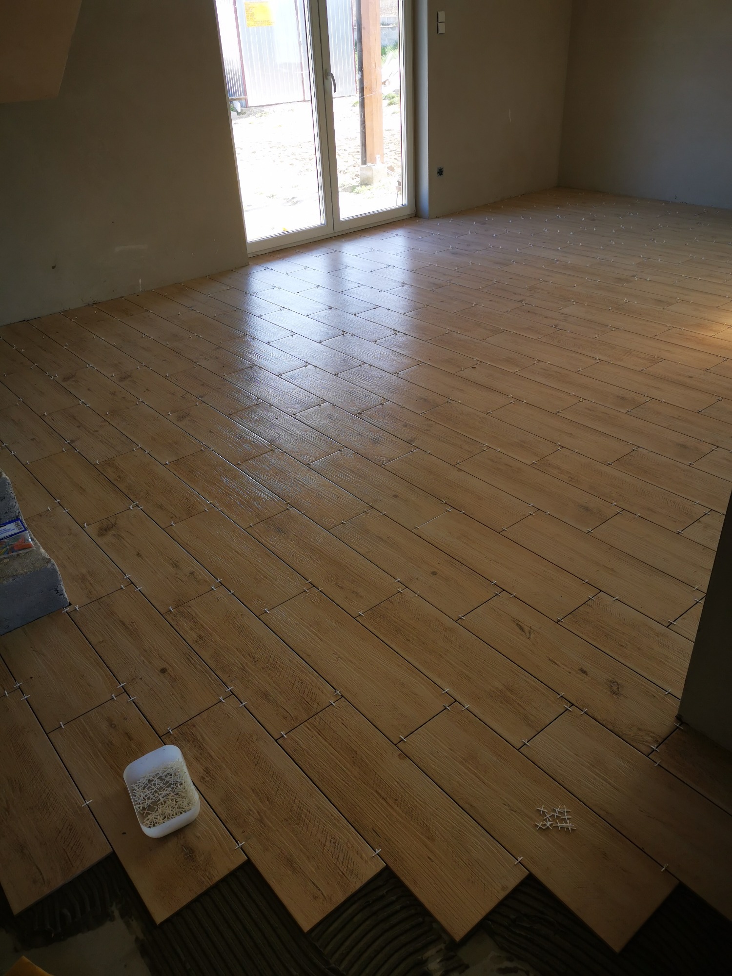 Układanie podłogi z płytek imitujących drewno w jasnym pomieszczeniu, widoczne krzyżyki dystansowe i pojemnik z nimi.