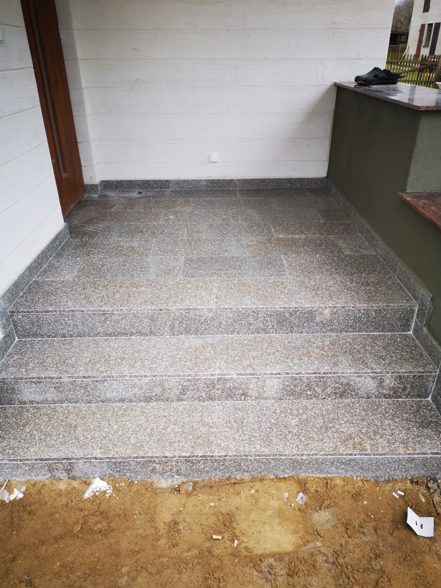 Granitowe schody wejściowe do domu, z widocznymi płytkami i fugami, częściowo ułożone, z ziemią i resztkami materiałów budowlanych u podstawy.