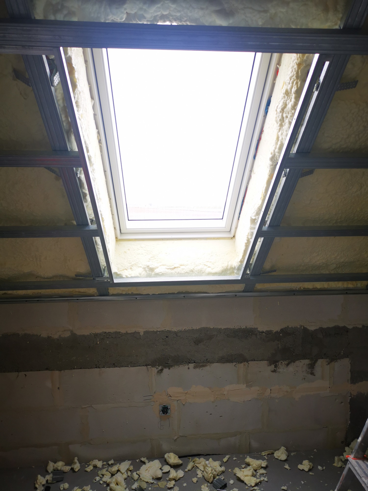 Poddasze w trakcie ocieplania pianką poliuretanową wokół okna dachowego, widoczna metalowa konstrukcja sufitu podwieszanego i fragment ściany z niedokończonymi pracami wykończeniowymi.