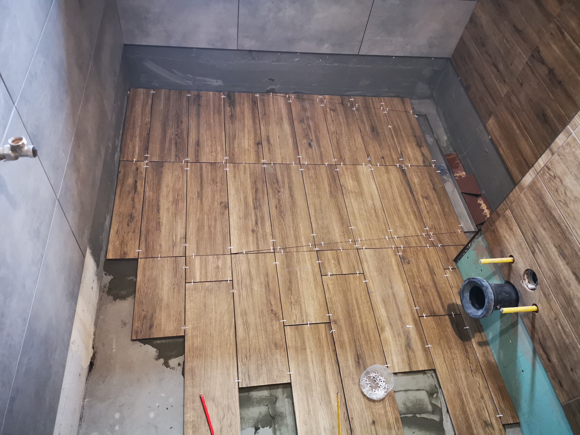 Montaż płytek drewnopodobnych w łazience, widoczne krzyżyki dystansowe, fragment instalacji kanalizacyjnej i narzędzia.
