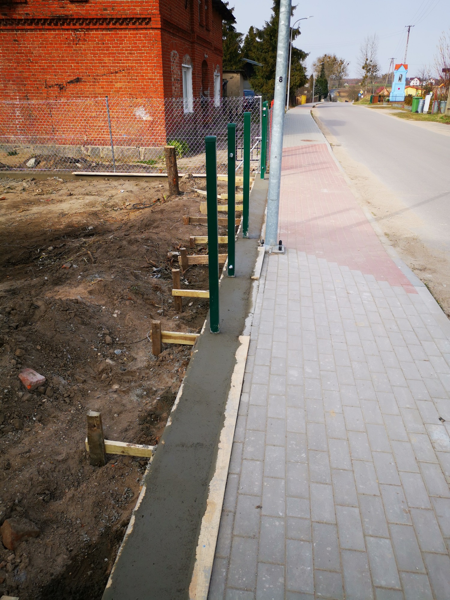 Świeżo wylany fundament pod ogrodzenie z zielonymi słupkami, widoczne drewniane szalunki i fragment chodnika z kostki brukowej przy ulicy.