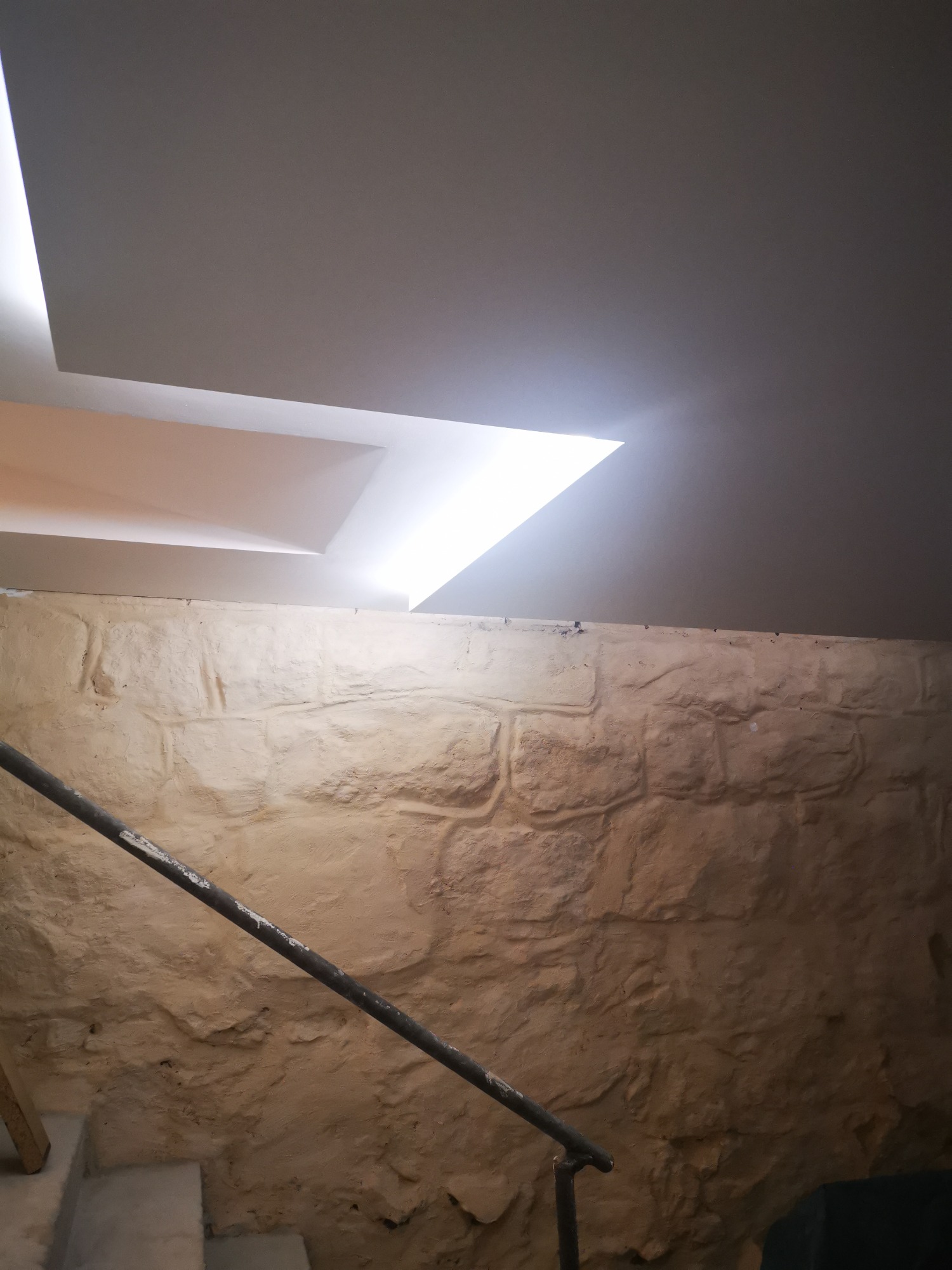 Surowa ściana z kamienia wapiennego zestawiona z nowoczesnym podwieszanym sufitem z oświetleniem LED, widok z dołu schodów.