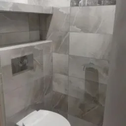 Nowoczesna toaleta w łazience wykończonej szarymi płytkami imitującymi marmur, z wbudowaną półką i przyciskiem spłukiwania w chromowanej ramce oraz dekoracyjnym panelem z motywem miejskim.