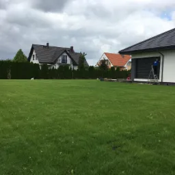 Rozległy trawnik przed domem z dwoma budynkami w tle, jeden z nich z ciemnym dachem i balkonem, a drugi z pomarańczowym dachem; w prawym dolnym rogu domu stoi pracownik na drabinie.