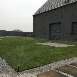 Nowoczesny ogród z trawnikiem, szarą elewacją domu i automatycznym nawadnianiem w trakcie pracy. W tle widoczny czarny płot i sąsiedni dom.