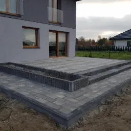 Nowoczesny taras z kostki brukowej w odcieniach szarości, z dwustopniowymi schodami i podwyższoną rabatą, przylegający do szarego domu z drewnianymi oknami i balkonem.