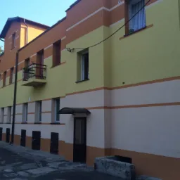 Odnowiona elewacja budynku wielorodzinnego z poziomymi pasami w kolorach żółtym, pomarańczowym i kremowym, widoczny balkon z czarną balustradą i doniczką z kwiatami.