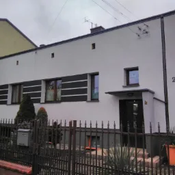 Dwukondygnacyjny dom z białą elewacją i poziomymi czarnymi pasami pod oknami, widoczny numer domu '2', ogrodzenie metalowe, pochmurne niebo.