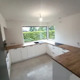 Jasna kuchnia z białymi szafkami i drewnianym blatem, widok na zielony ogród przez duże okno, minimalistyczne oświetlenie.