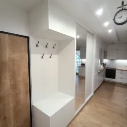 Biały przedpokój z szafą wnękową z lustrzanymi drzwiami przesuwnymi, wbudowanym siedziskiem z szafkami i wieszakami, widok na kuchnię w tle.