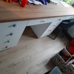 Biurko dziecięce z blatem imitującym drewno i białymi szafkami z szufladami, których uchwyty mają kształt piłek do różnych sportów, na blacie zabawki i przybory szkolne, obok pojemniki z zabawkami.