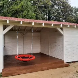 Biała drewniana altana z czerwonym dachem i huśtawką typu gniazdo zawieszoną wewnątrz, widok na zielony ogród w tle.