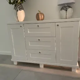 Biała komoda z trzema szufladami i dwojgiem drzwiczek, z dekoracjami na blacie w postaci wazonu i lampy, ustawiona na szarych płytkach.