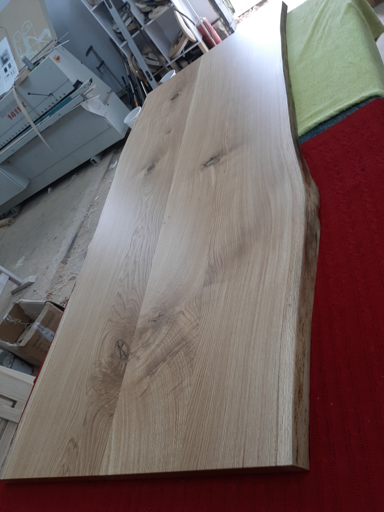 Surowy blat drewniany z naturalną krawędzią, leżący w warsztacie na czerwonej tkaninie, w tle maszyna stolarska i półki z materiałami.