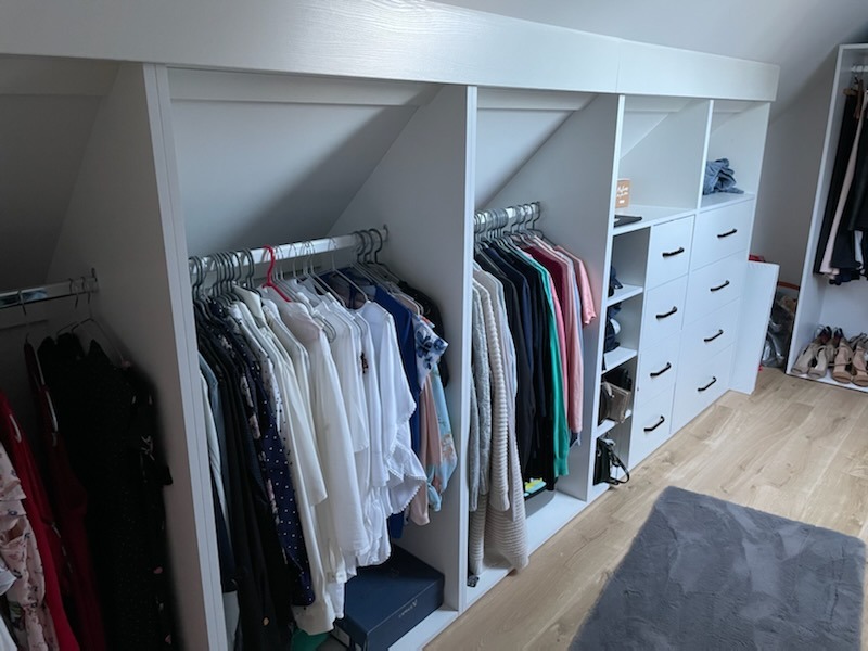 Biała, zabudowana szafa wnękowa na poddaszu ze skośnym sufitem, z ubraniami na wieszakach i szufladami, uzupełniona o półki na buty i akcesoria.