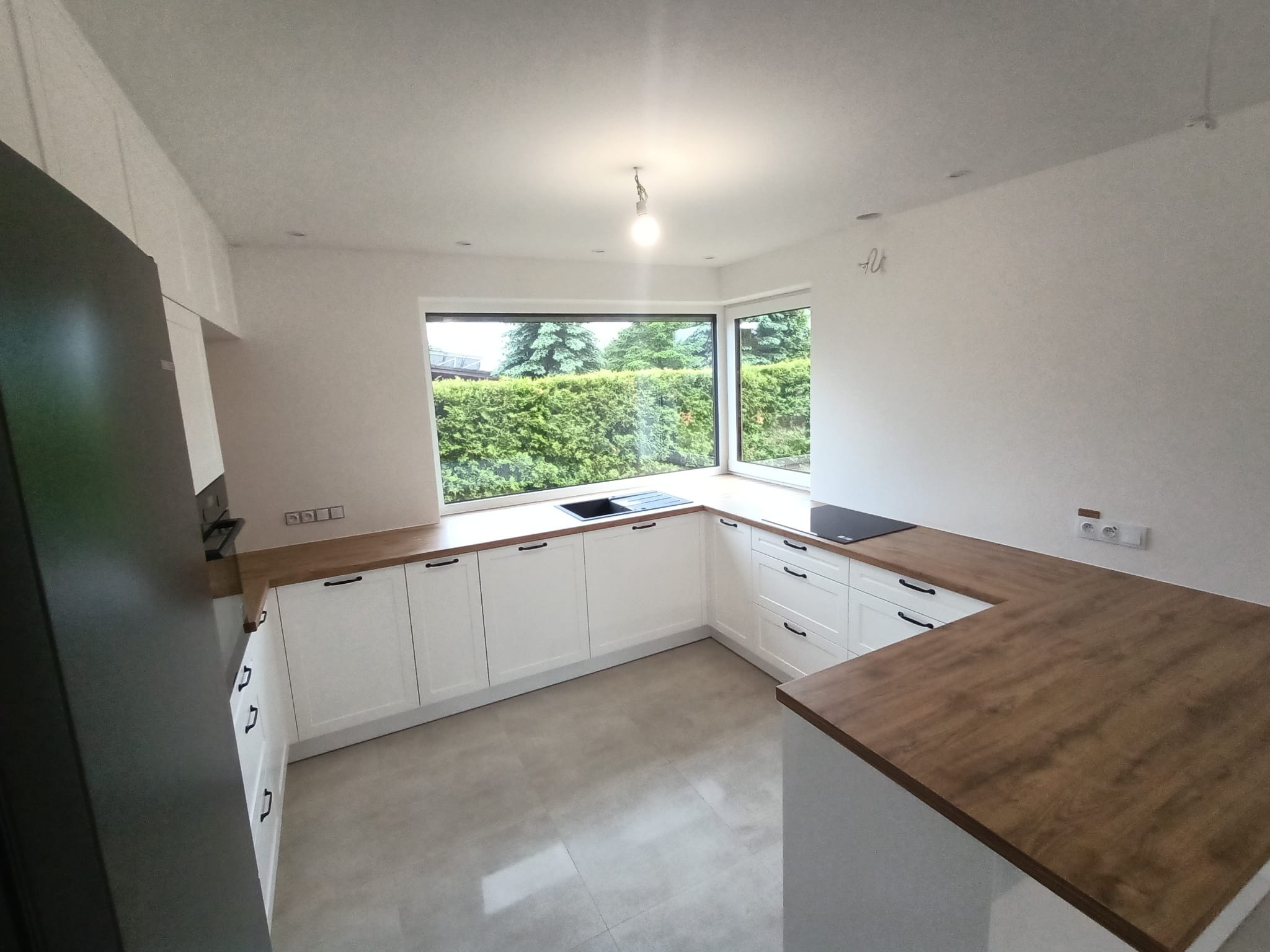 Jasna kuchnia z białymi szafkami i drewnianym blatem, widok na zielony ogród przez duże okno, minimalistyczne oświetlenie.