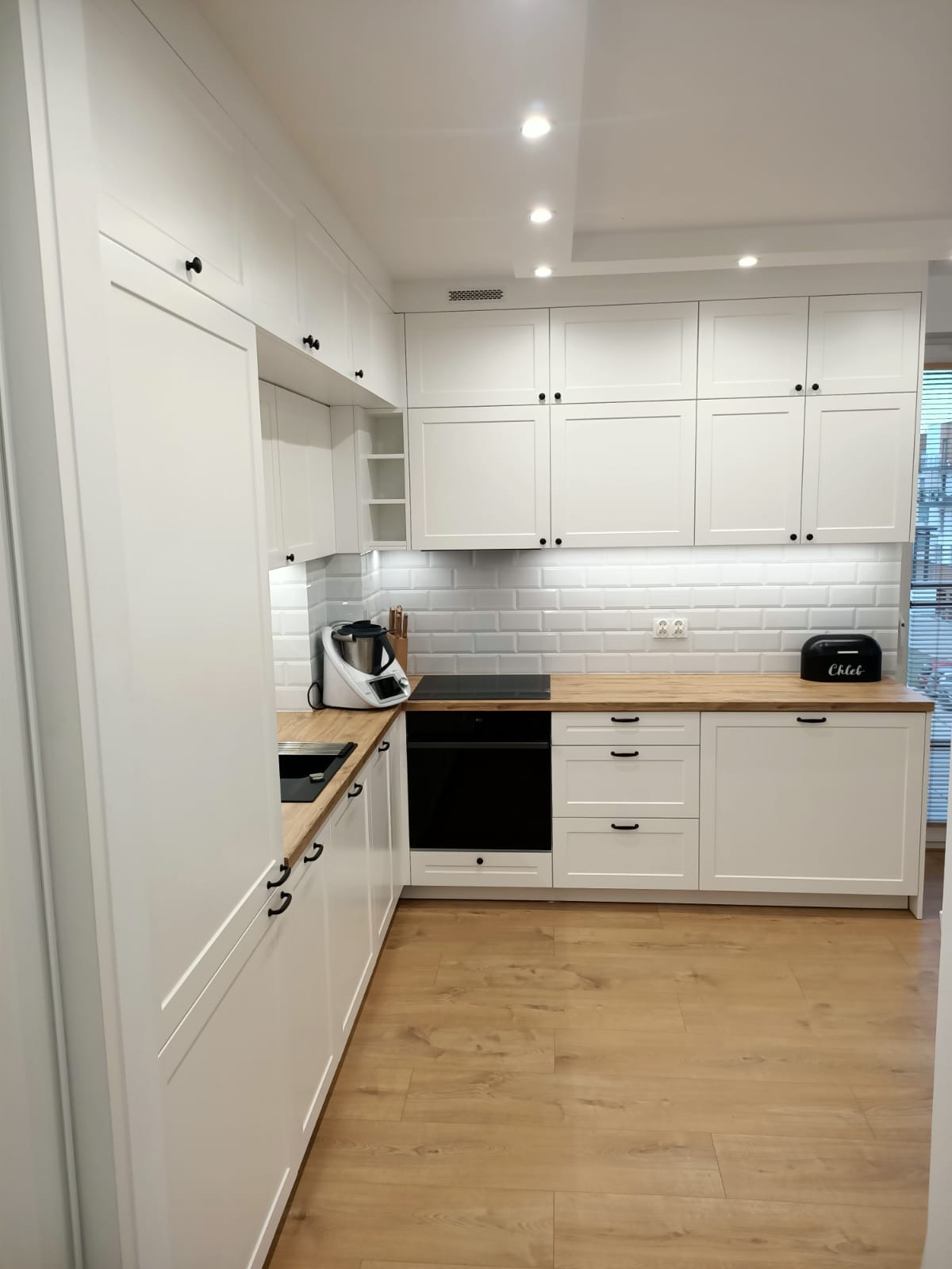 Nowoczesna, jasna kuchnia z białymi szafkami z czarnymi uchwytami, drewnianym blatem i białymi płytkami imitującymi cegłę, wyposażona w czarny piekarnik, płytę indukcyjną, robot kuchenny i chlebak.