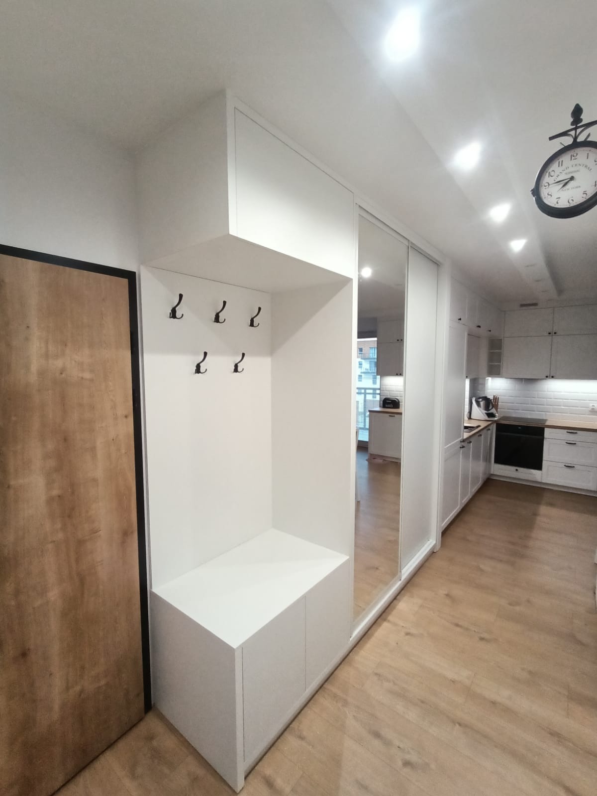 Biały przedpokój z szafą wnękową z lustrzanymi drzwiami przesuwnymi, wbudowanym siedziskiem z szafkami i wieszakami, widok na kuchnię w tle.